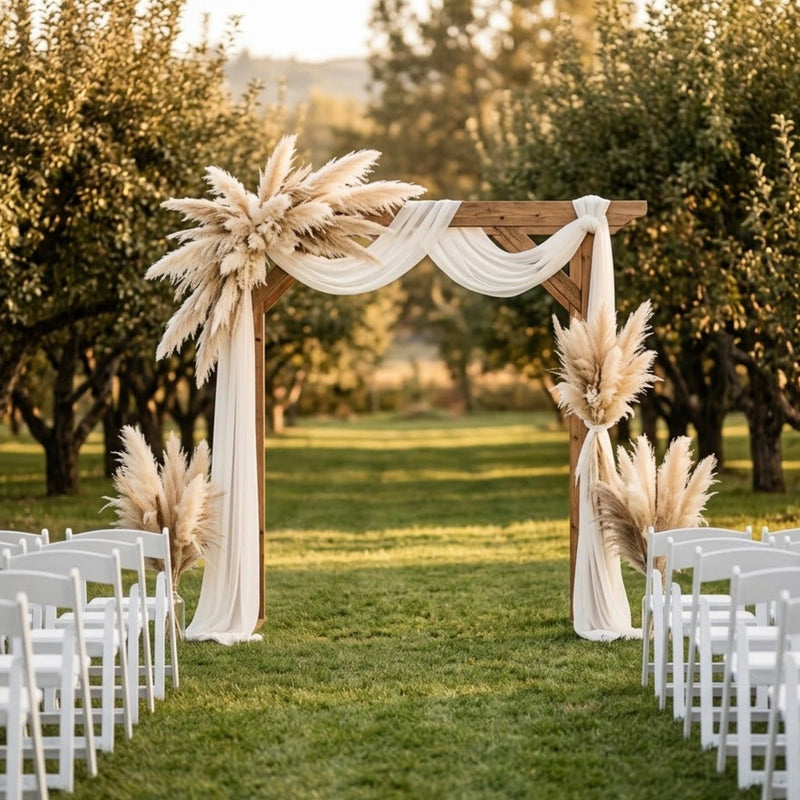 Arche de mariage en bois ornée de plumes de pampa artificielle et de voilage blanc, au coucher du soleil