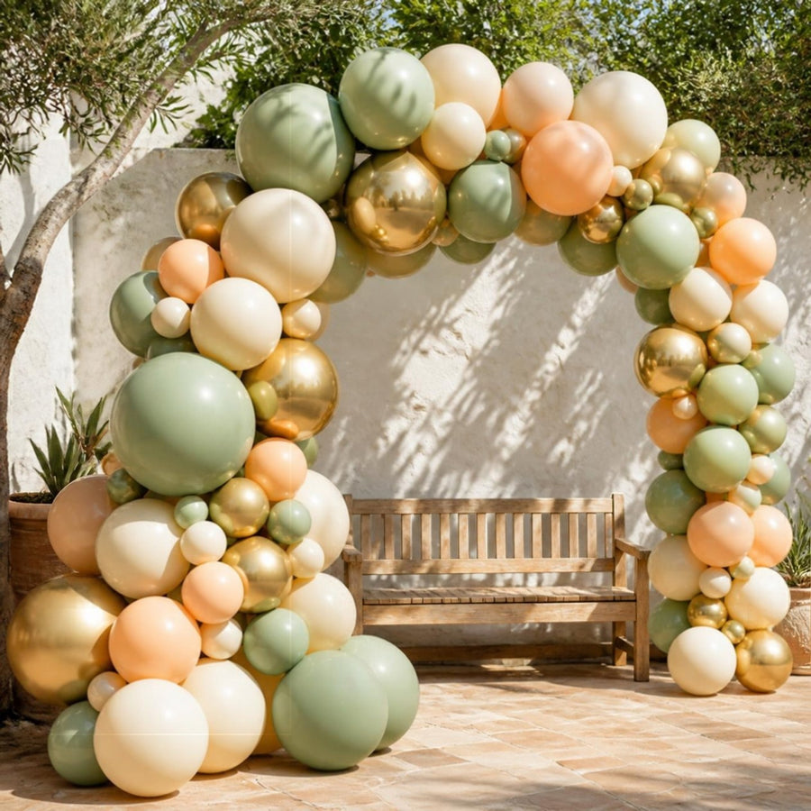 Arche de ballons d'anniversaire sauge, crème, pêche et or contre un mur blanchi sur une terrasse ensoleillée