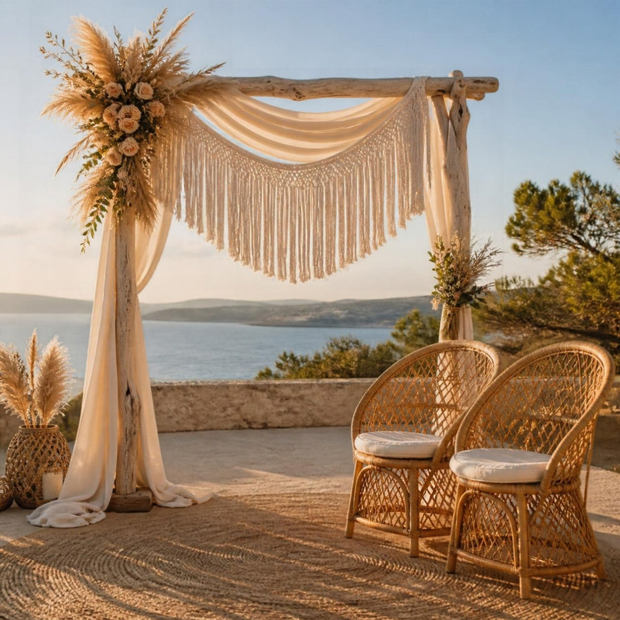 Arche de mariage bohème en bois clair avec tissu crème et franges, tapis tissé sable et chaises en rotin sur une terrasse