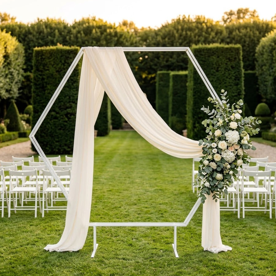 Arche de mariage hexagonale en métal blanc mat sur pieds, voilage ivoire et fleurs ivoire et eucalyptus, dans un jardin à la française