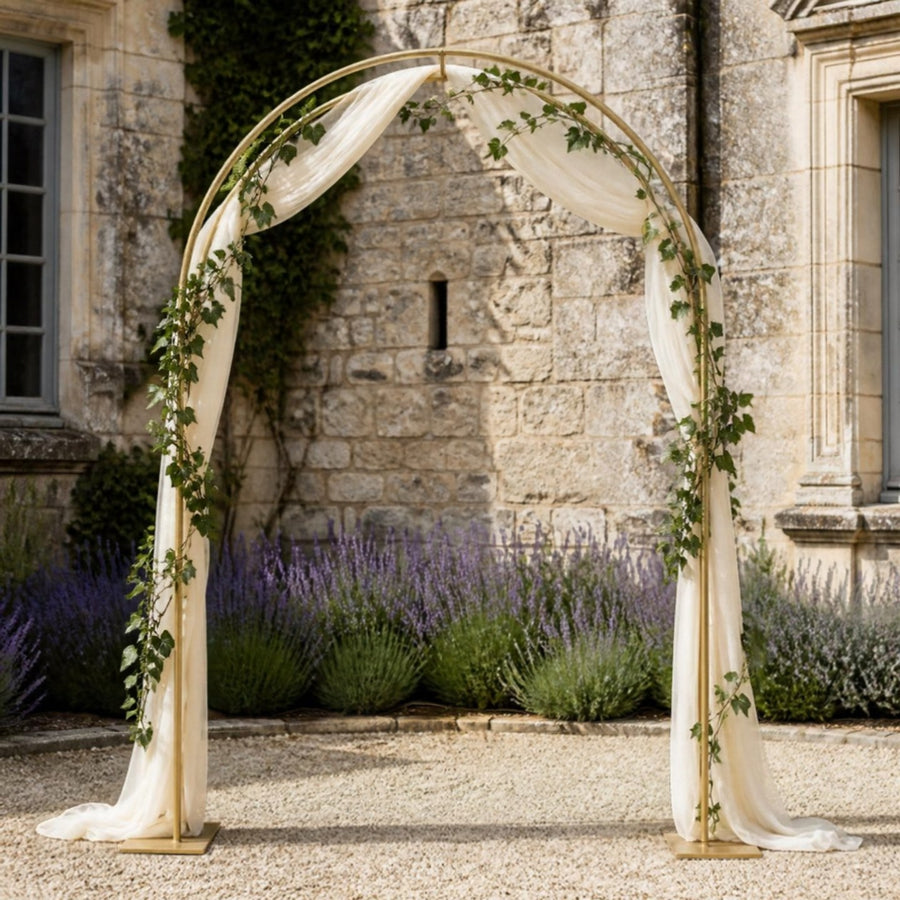 Arche de mariage en métal doré champagne à sommet arrondi, voile ivoire et lierre, dans une cour devant un manoir