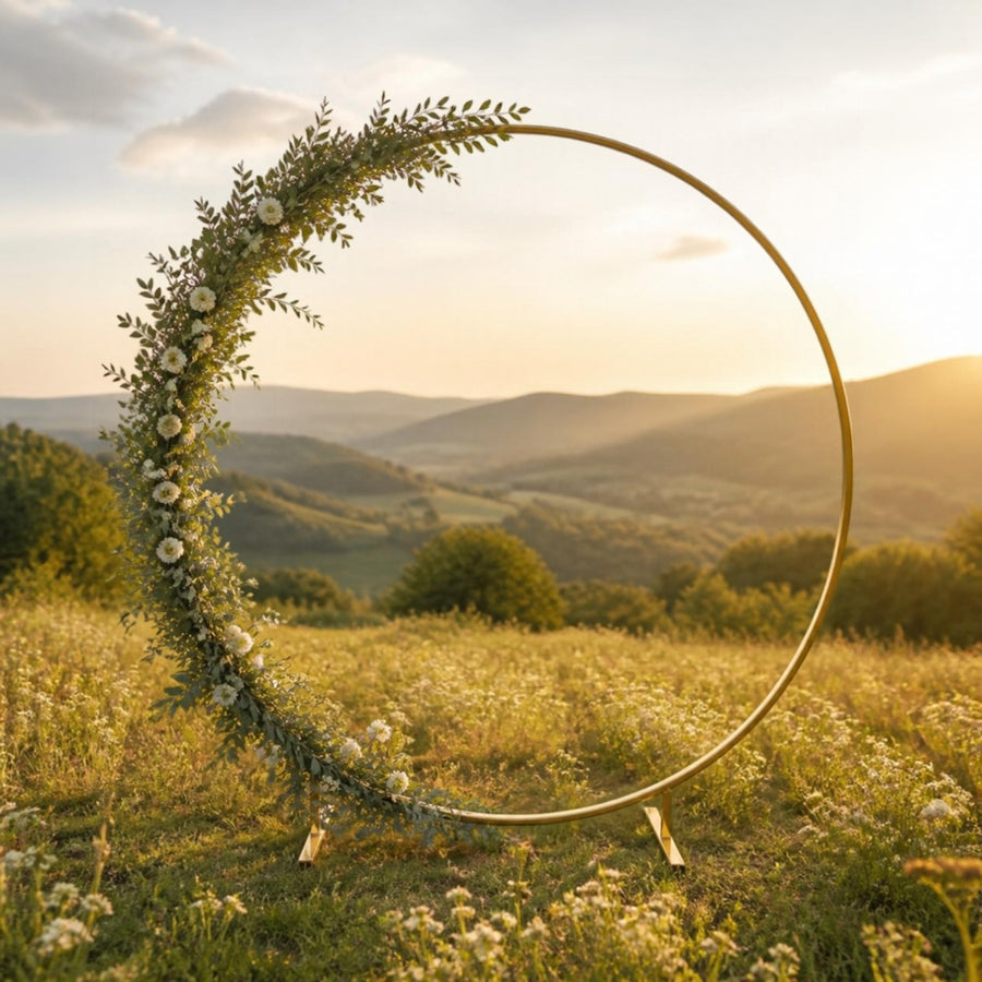 Grande arche de mariage ronde dorée ornée d'une guirlande de verdure sauge et de fleurs ivoire dans une prairie fleurie