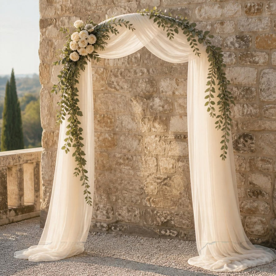 Arche de mariage décorée d'un voilage ivoire, d'une guirlande de feuillage sauge et d'un bouquet de roses, sur une terrasse