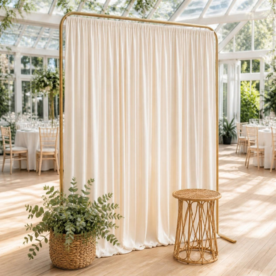 Fond de photobooth en tissu plissé ivoire sur support doré, avec tabouret en rotin, dans une salle lumineuse type serre