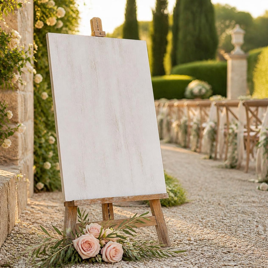 Panneau de bienvenue en bois blanchi sur chevalet rustique à l'entrée d'un jardin, avec roses pâles et branche d'olivier